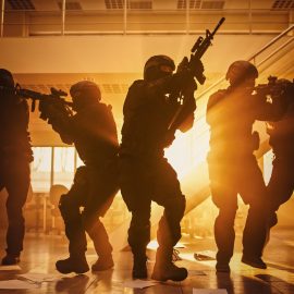 Masked Fireteam of Armed SWAT Police Officers Storm a Sunny Seized Office Building with Desks and Computers. Soldiers with Rifles Move Forwards and Cover Surroundings. Shot with Yellow Warm Filter.